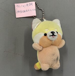 ボールチェーンが付いた犬のぬいぐるみの写真です。ボールチェーンは、ぬいぐるみの頭の部分についた輪に通されています。ぬいぐるみの色は、体が薄い黄色で、顔の下部分からお腹が薄い茶色です。尻尾は薄い黄色と薄い茶色の縞模様です。